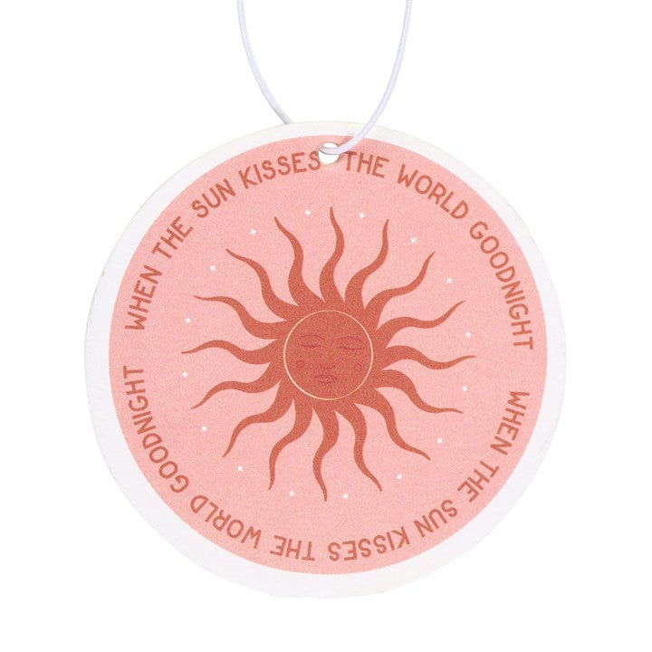 Close up image of a pink and gold round car airfreshener depicting the sun with the phrase when the sun kisses the world goodnight.