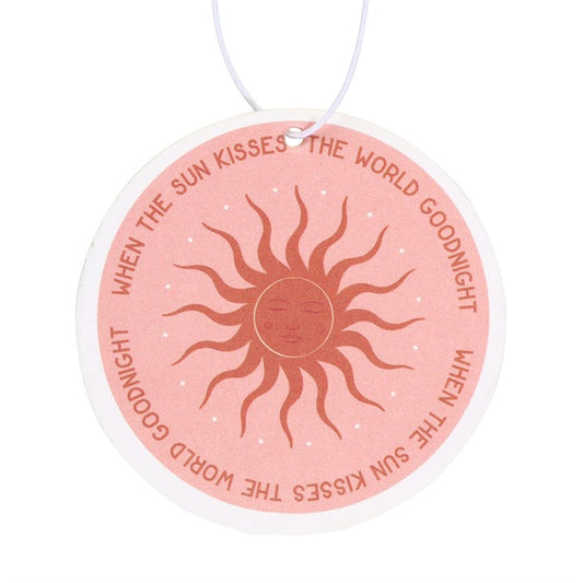 Close up image of a pink and gold round car airfreshener depicting the sun with the phrase when the sun kisses the world goodnight.
