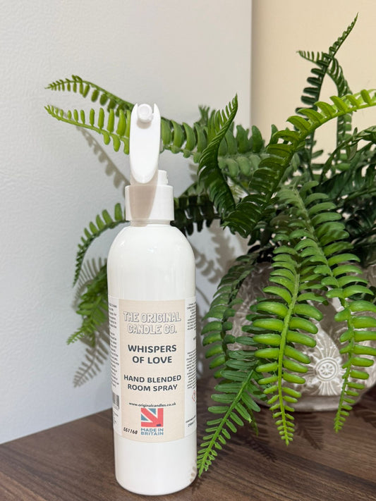 White room spray bottle labeled 'Whispers of Love' on a wooden surface with a fern plant in the background.